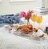 Clear acrylic serving tray with sides on a bed displaying a small vase of fresh cut flowers, 2 stemmed orange juice glasses, plates of muffins, scones, fruit and coffee.