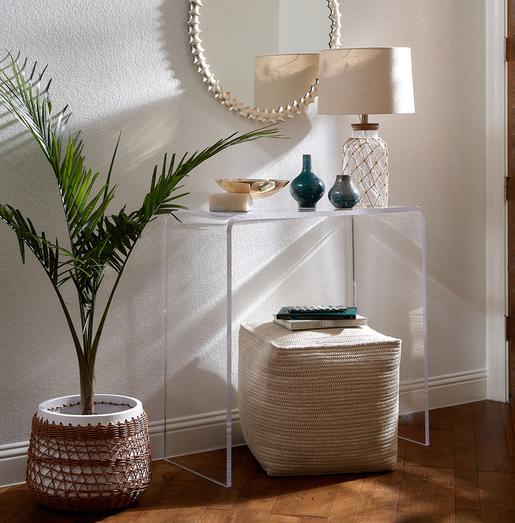 Clear acrylic waterfall edge entry or hallway table displaying a lamp, decorative vases and bowl with pouf underneath against a wall next to a tall plant in an entryway.