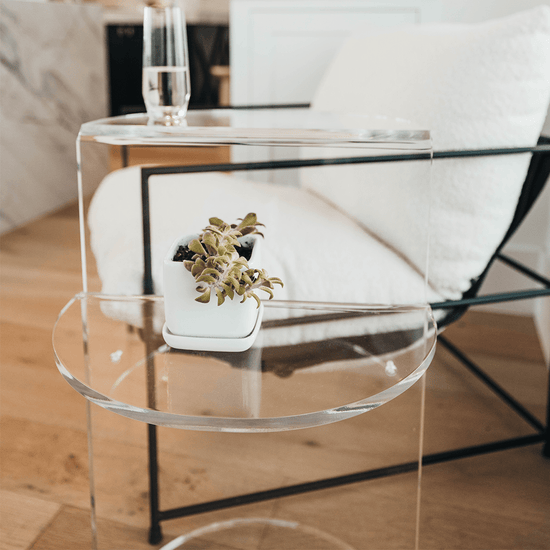 Close up of a clear acrylic side table featuring a top shelf extending 1 direction and the second shelf extending the opposite direction displaying a glass of water and a plant beside a chair.