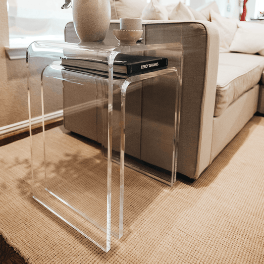 2 clear acrylic waterfall edge nesting end tables with book, vase, and decor next to a couch in a living room.