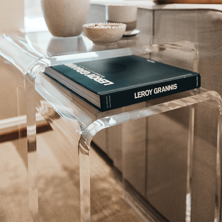 2 clear acrylic waterfall edge nesting end tables with book, vase, and decor next to a couch in a living room with the lower table pulled out.