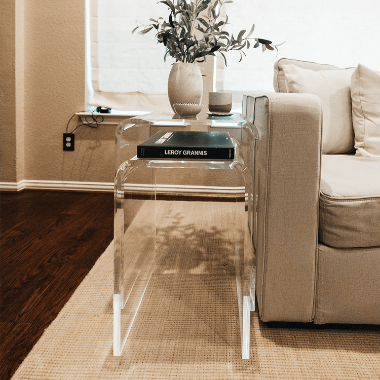 2 clear acrylic waterfall edge nesting end tables with book, vase, and decor next to a couch in a living room with the lower table pulled out.