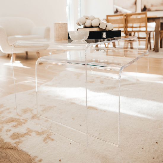 2 clear acrylic waterfall edge nesting tables featuring the larger 20" top table displaying books, candle, and decor and the smaller table is pulled slightly out. The tables are placed in a living room on an area rug.