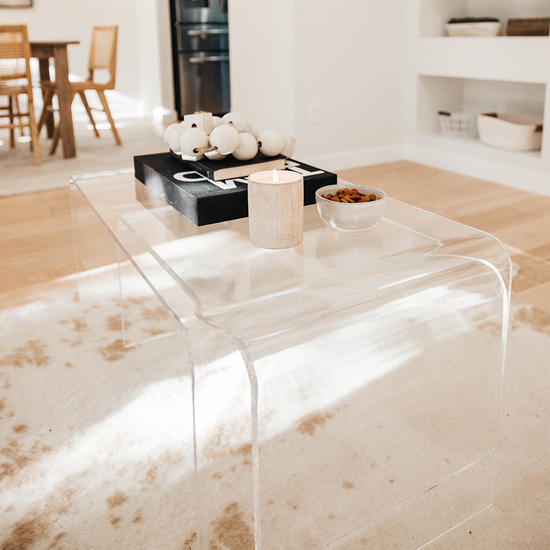 2 clear acrylic waterfall edge nesting tables featuring the larger 20" top table displaying books, candle, and decor and the smaller table is tucked underneath. The tables are placed in a living room on an area rug.