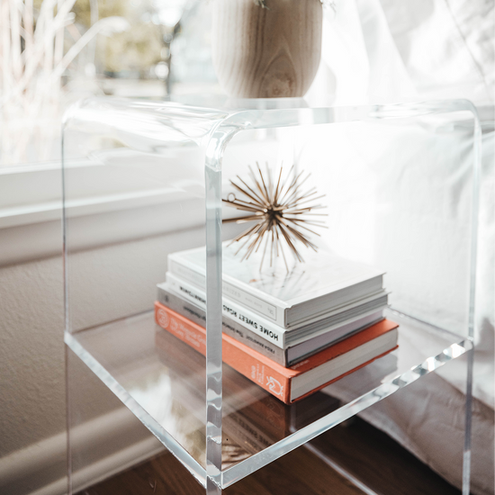 Closeup of a clear acrylic nightstand displaying books, decor, and vase beside a bed and a window.