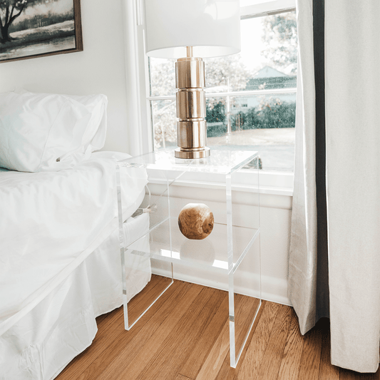 Side angle of a clear acrylic slab nightstand with a lamp placed on top and 1 interior shelf in front of a window next to a bed.