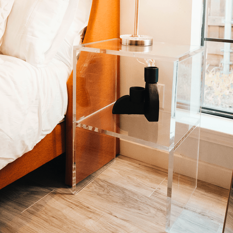 Clear acrylic slab nightstand with a lamp placed on top and 1 interior shelf displaying art decor in front of a window next to a bed.