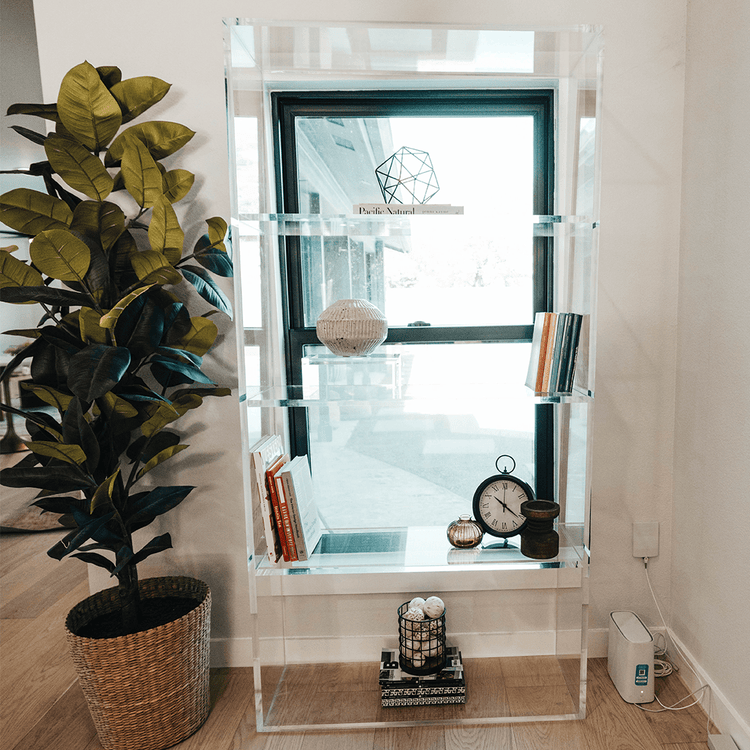 Vertical clear acrylic 5 interior shelf bookcase decorated with a clock, books, and decor next to a tall plant in front of a window showing a view outside..