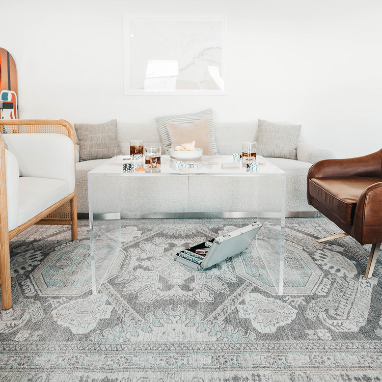 Clear acrylic 23" slab coffee table displaying drink glasses, snacks, and a game centered on an area rug between 2 chairs and a couch.