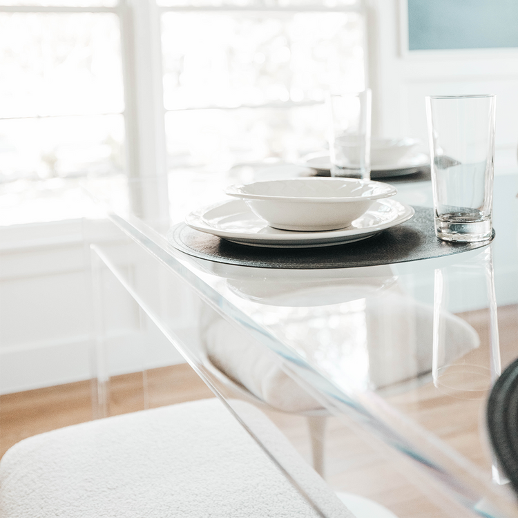 Closeup of the edge on a clear acrylic dining room table displaying glasses and place settings with views of the chairs underneath the table.