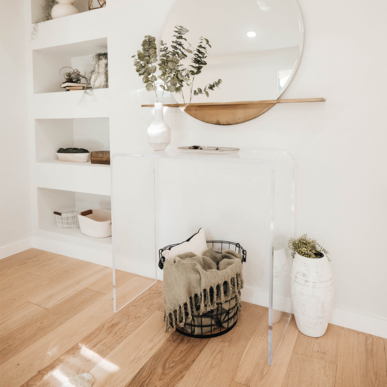Clear acrylic waterfall edge entry or hallway table displaying a decorative vase and plate with a pillow and throw basket underneath against a wall in living room.